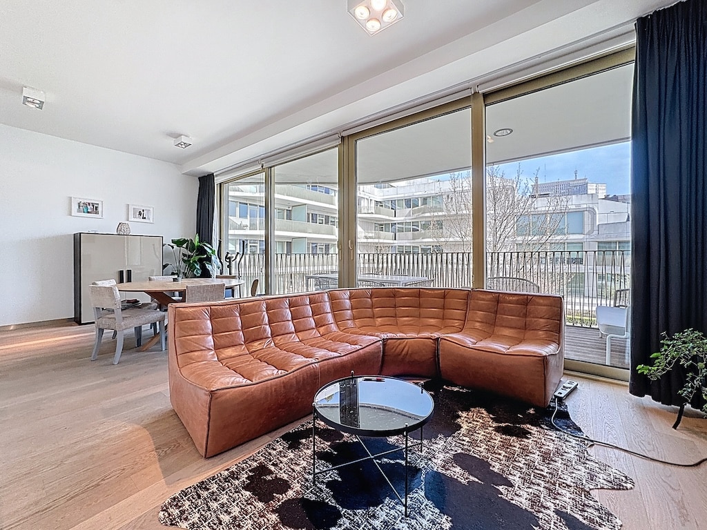 Salon contemporain lumineux avec canapé en cuir, grandes fenêtres, table en verre et espace repas élégant.