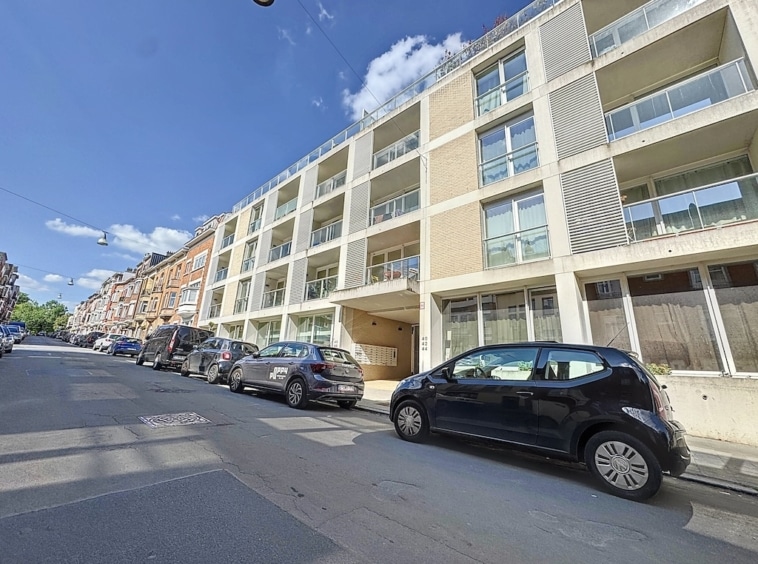 Apartment moderne avec grandes fenêtres et balcons en centre-ville animé, sous ciel bleu ensoleillé.