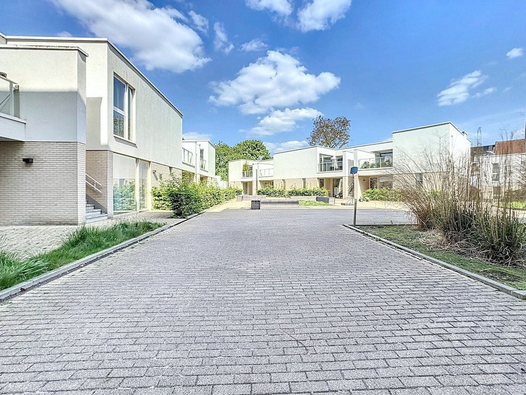 Environnement moderne avec bâtiments lumineux, chemin pavé et verdure sous ciel partiellement nuageux.