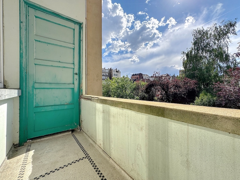 Balcon ensoleillé avec porte turquoise, vue verdoyante et ciel nuageux, ambiance paisible et charmante.