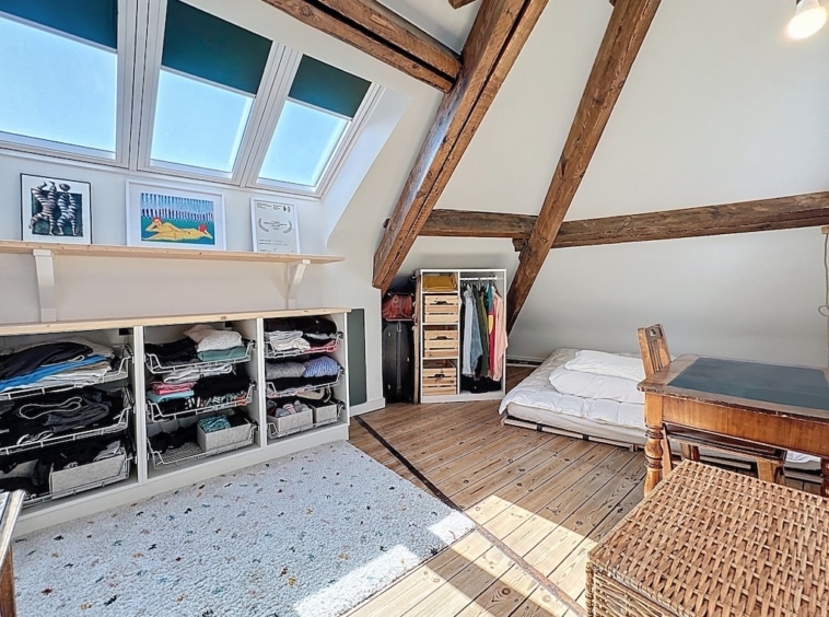 Chambre d’attique lumineuse, cosy, avec poutres en bois, rangement organisé et lumière naturelle abondante.