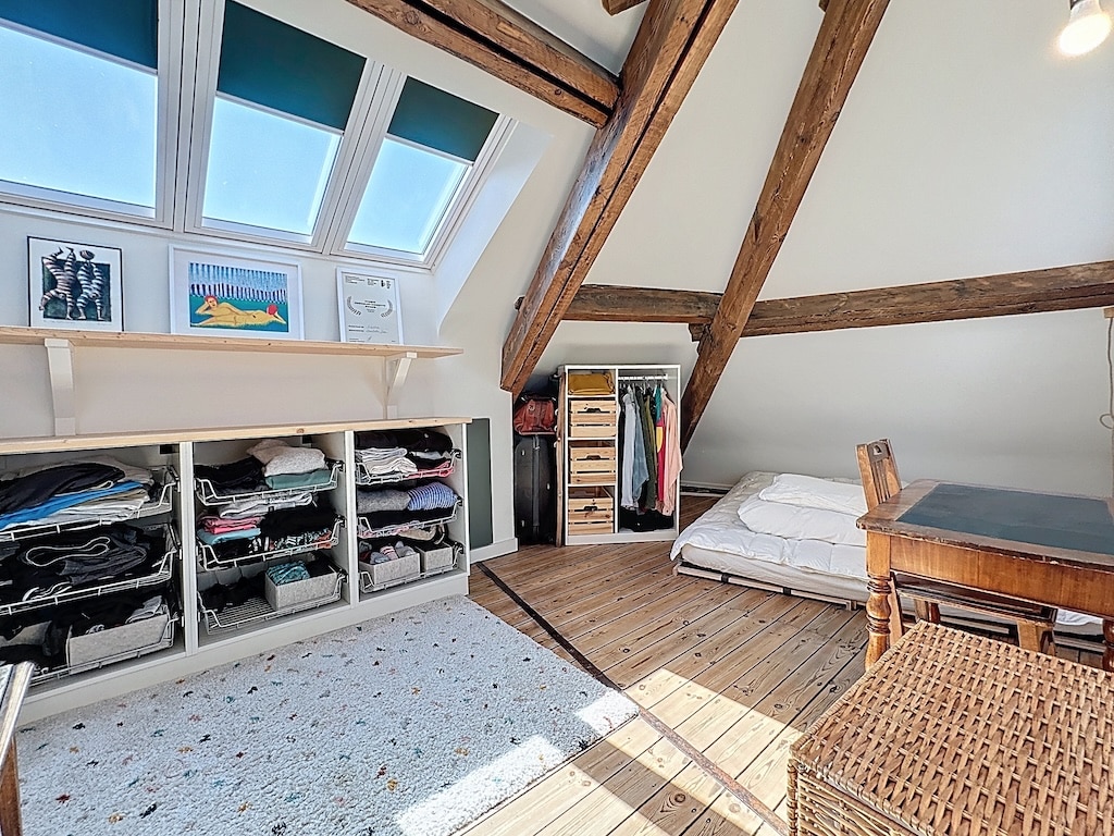 Chambre d’attique lumineuse, cosy, avec poutres en bois, rangement organisé et lumière naturelle abondante.
