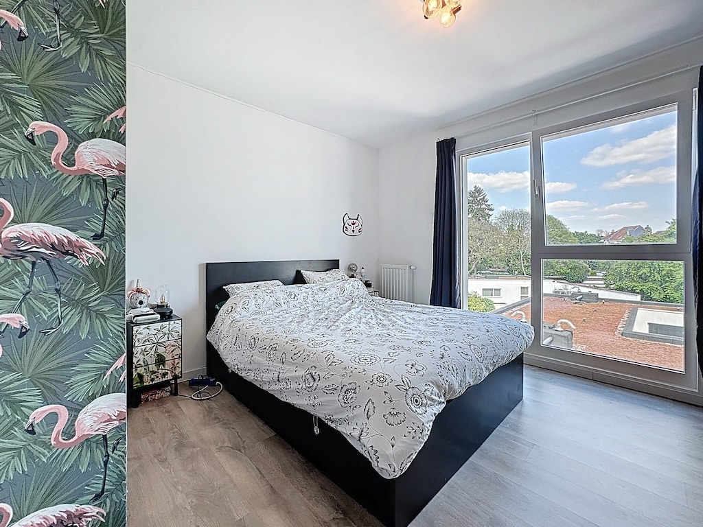 Chambre lumineuse moderne avec mur tropical flamants roses, lit blanc et vue verdoyante.