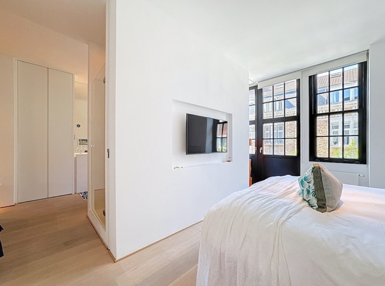 Chambre minimaliste lumineuse avec lit blanc, grande fenêtre, mur TV encastré et sol en bois chaleureux.