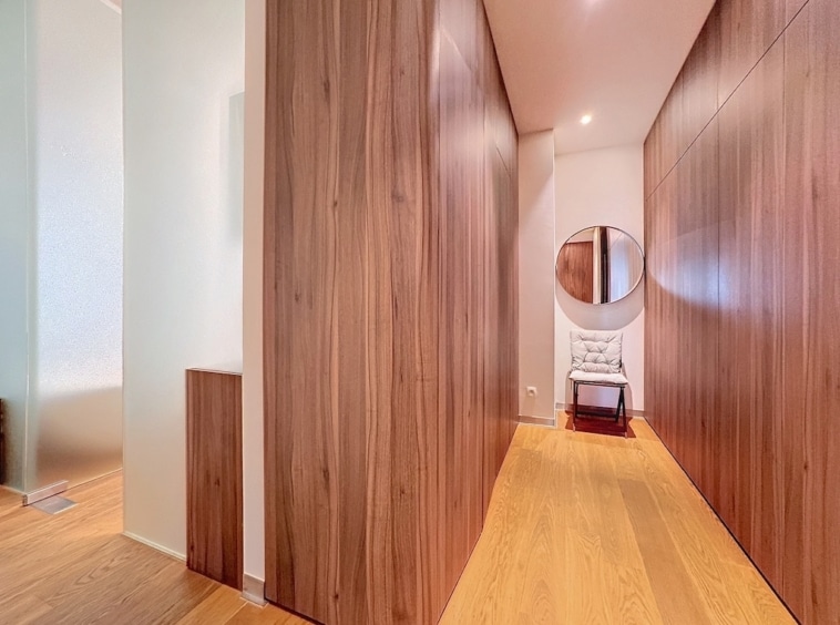 Couloir moderne en bois, miroir circulaire, fauteuil clair, ambiance chaleureuse et lumineuse.
