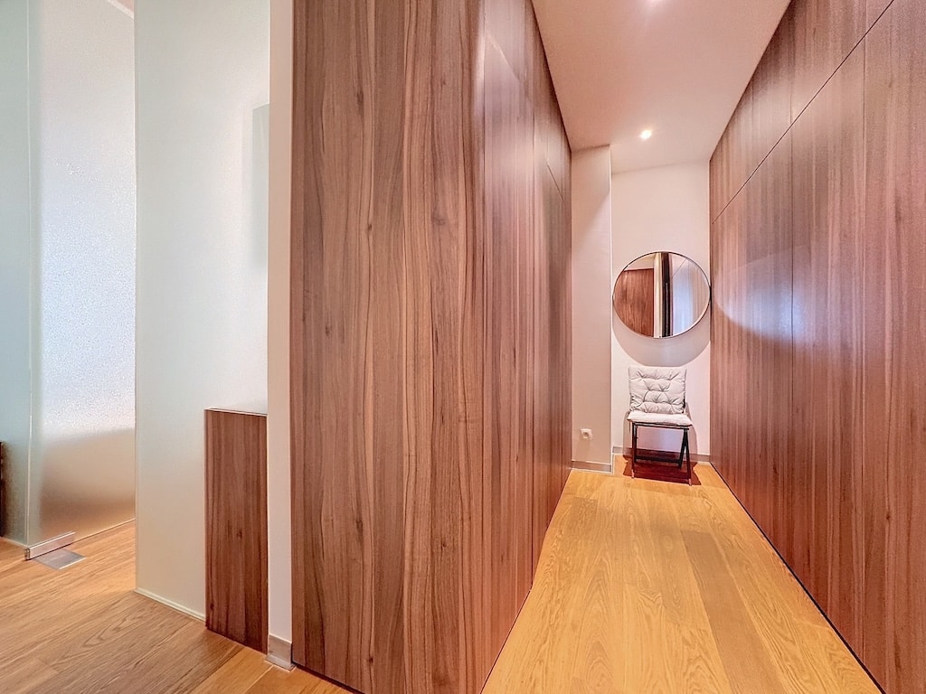 Couloir moderne en bois, miroir circulaire, fauteuil clair, ambiance chaleureuse et lumineuse.