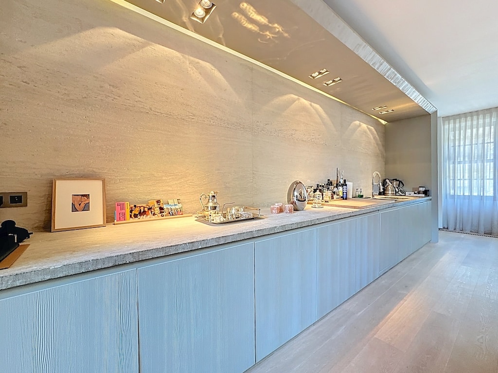 Cuisine moderne minimaliste avec plan de travail en pierre, éclairage chaud et grande fenêtre lumineuse.