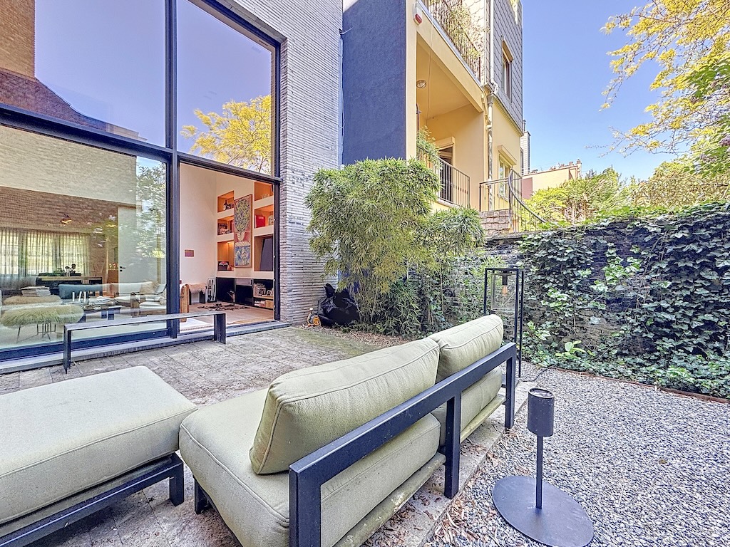 Espace extérieur moderne avec canapés, éclairage d’ambiance, jardin urbain et façade en verre.