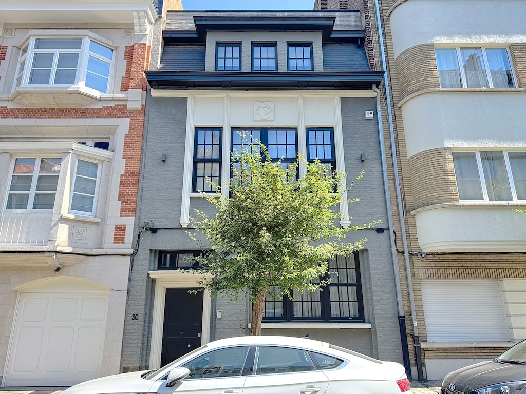 Maison moderne avec façade grise, fenêtres noires, porte centrale, arbre ornemental et voiture blanche.