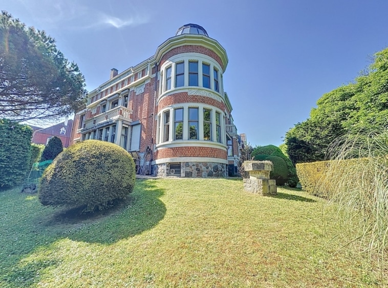 Manoir en briques rouges avec dôme, jardins soignés et arbres majestueux sous ciel bleu.
