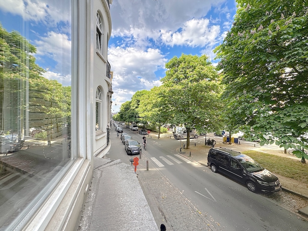 Rue pittoresque avec arbres, voitures et piéton en rouge sous un ciel bleu nuageux.