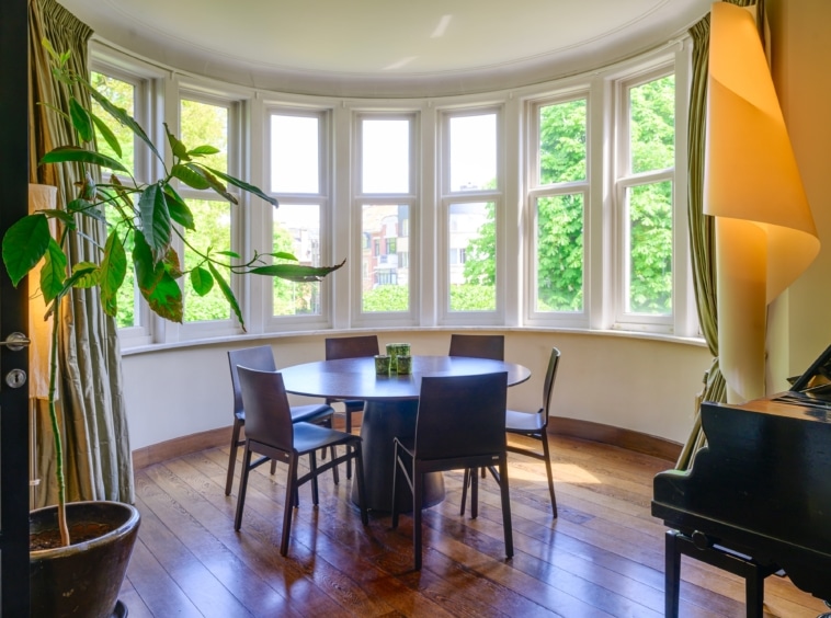 Salle à manger lumineuse avec vue verdoyante, piano élégant, table ronde et plantes vertes.