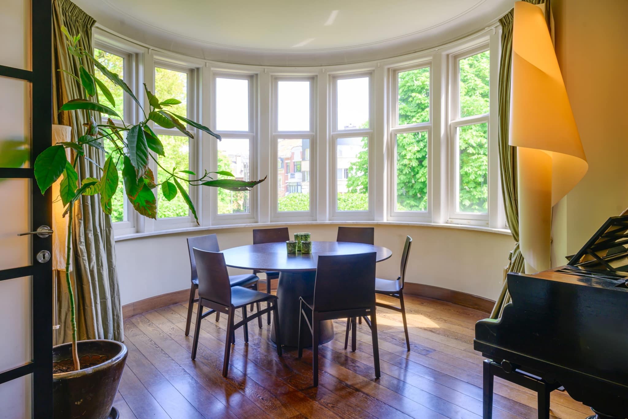 Salle à manger lumineuse avec vue verdoyante, piano élégant, table ronde et plantes vertes.