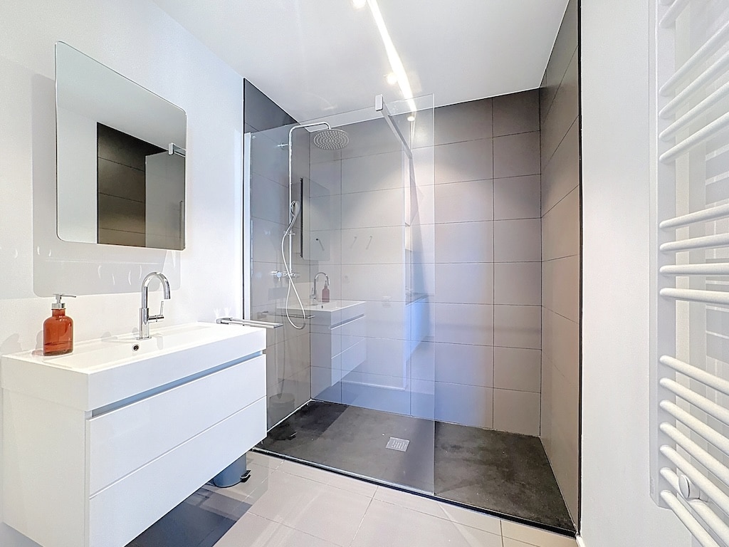 Salle de bain moderne, élégante, lumineuse avec douche à l’italienne, miroir carré et radiateur design.