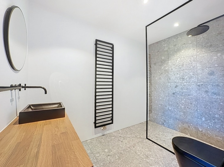 Salle de bain moderne minimaliste avec douche en verre, miroir rond et radiateur noir vertical.