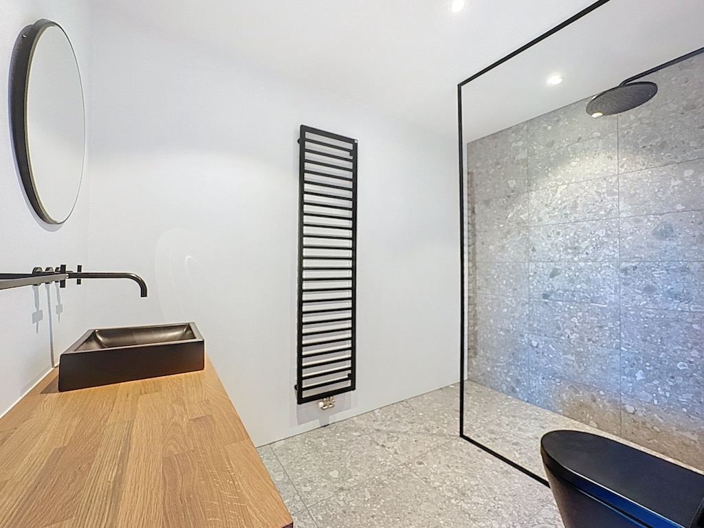 Salle de bain moderne minimaliste avec douche en verre, miroir rond et radiateur noir vertical.