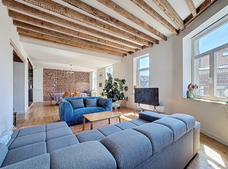 Salon moderne avec poutres en bois, canapé gris, fauteuil coloré, mur en briques et ambiance chaleureuse.