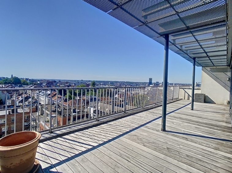 Balcon en bois avec vue sur la ville, structure ombragée et pot de fleurs, journée ensoleillée.