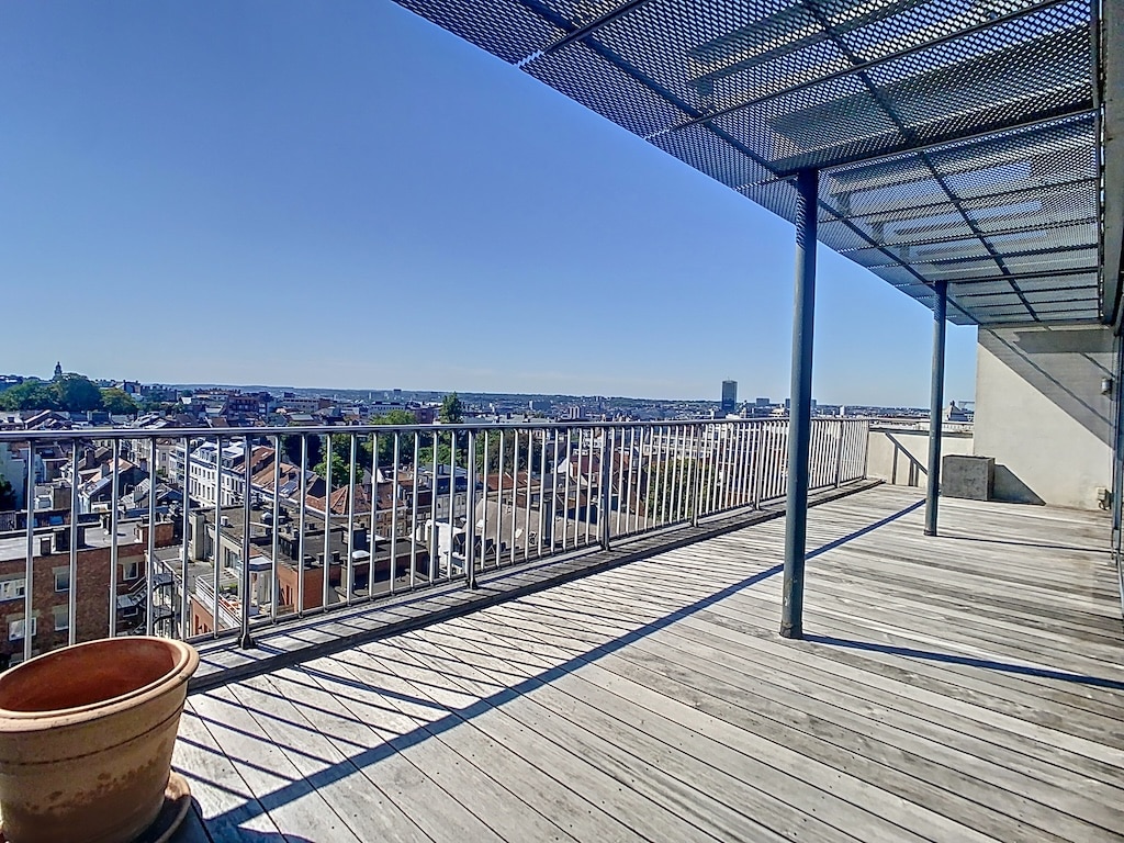 Balcon en bois avec vue sur la ville, structure ombragée et pot de fleurs, journée ensoleillée.