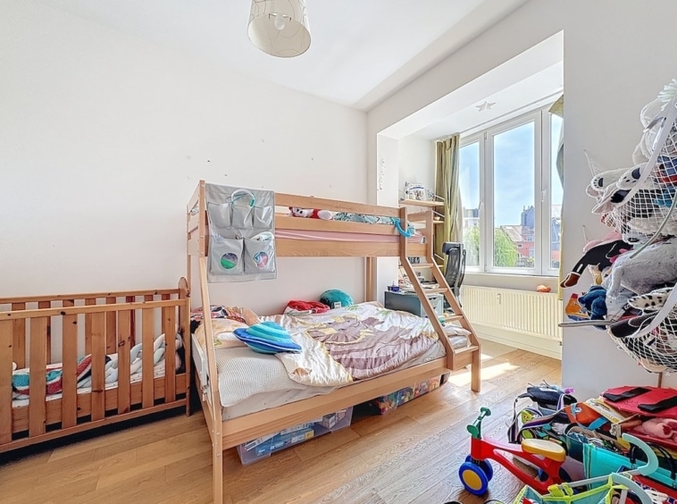 Chambre enfant chaleureuse avec lit superposé, berceau, jouets colorés et grande fenêtre lumineuse.