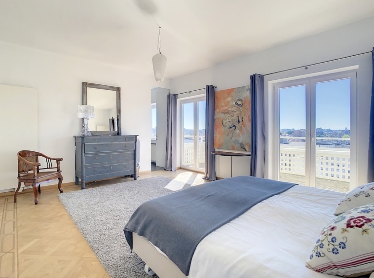 Chambre moderne lumineuse avec vue, lit élégant, mobilier sobre et coin lecture chaleureux.