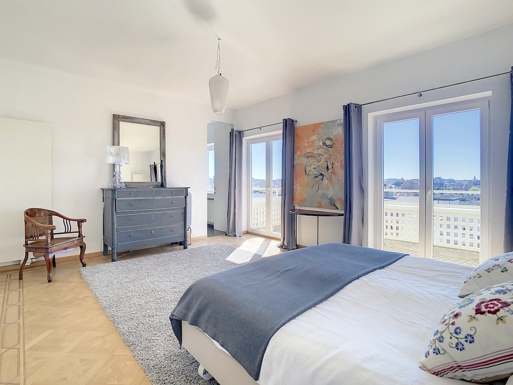 Chambre moderne lumineuse avec vue, lit élégant, mobilier sobre et coin lecture chaleureux.