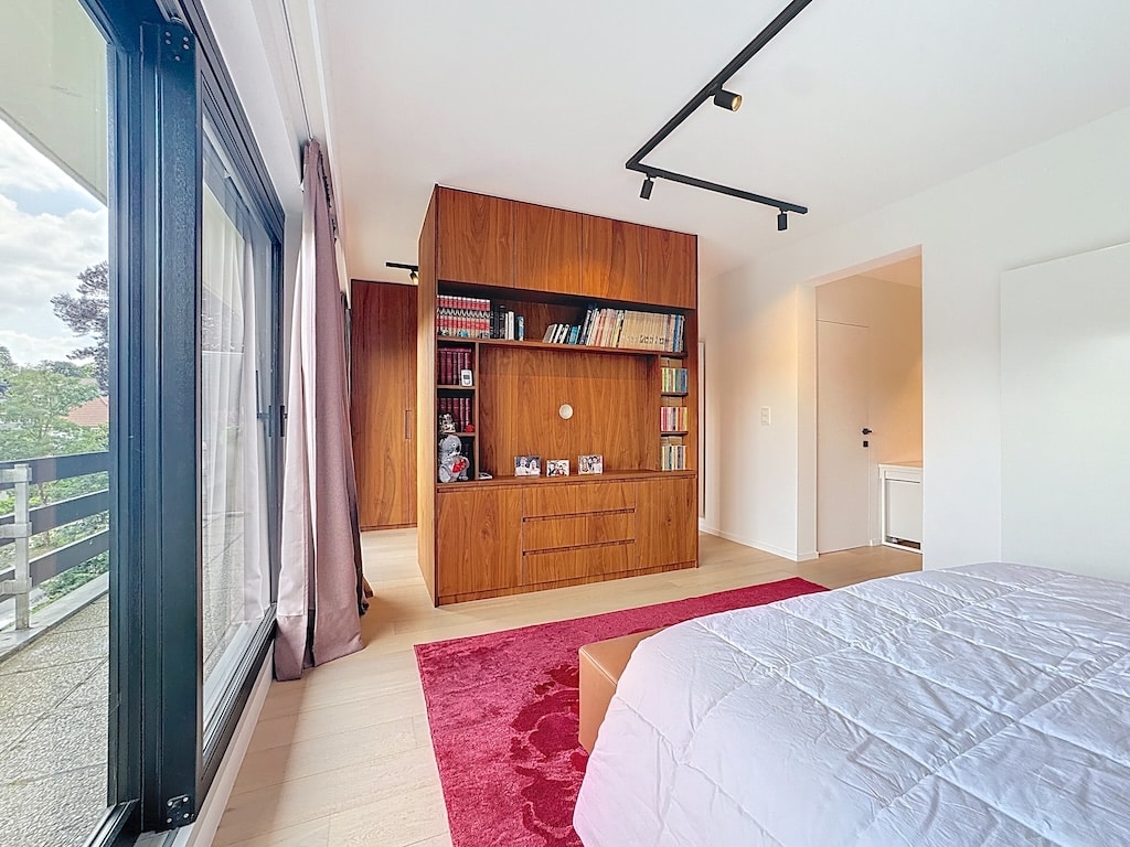 Chambre moderne lumineuse avec grande fenêtre, vue nature, lit épuré, étagère en bois et éclairage minimaliste.