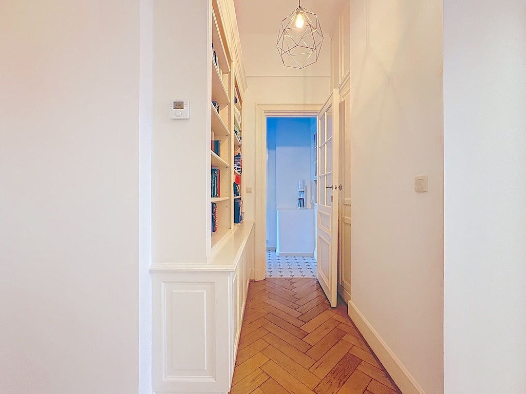 Couloir moderne avec bibliothèque intégrée, éclairage design, sol en bois chevron, élégance minimaliste.