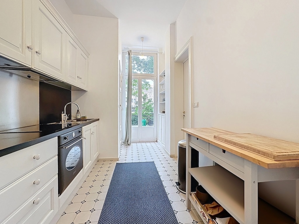 Cuisine moderne lumineuse avec îlot en bois, plan noir, large fenêtres et sol élégant en carreaux.