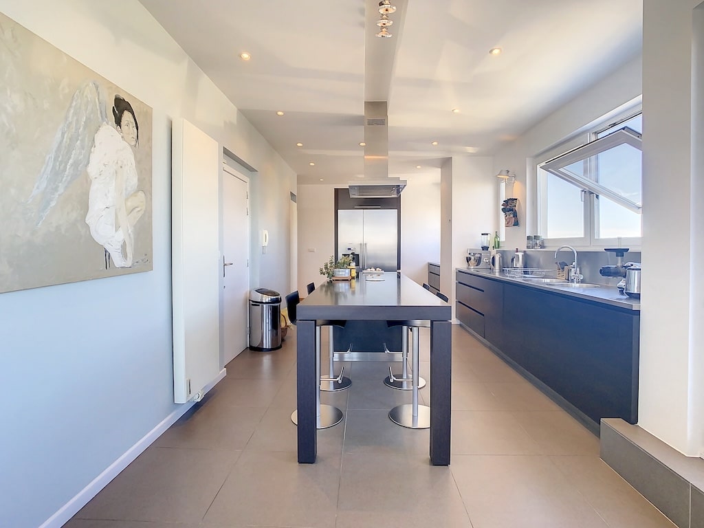 Cuisine contemporaine lumineuse avec îlot central, meubles sombres et grandes fenêtres naturelles.