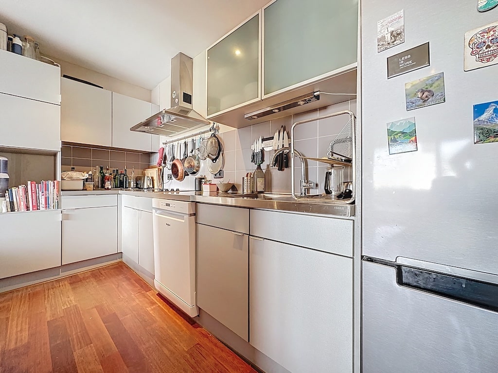 Cuisine moderne blanche avec plans en acier, réfrigérateur magnétique, hotte en métal, sol en bois.