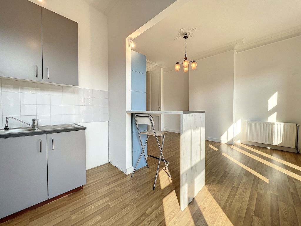 Cuisine moderne lumineuse avec rangements gris, espace repas, lumière suspendue et sol en bois clair.