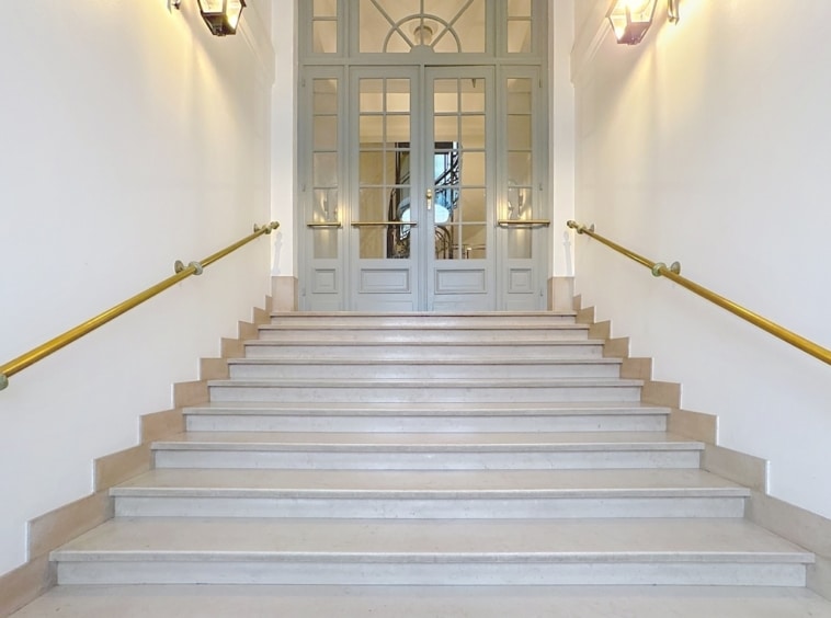 Escalier en pierre clair avec rampes en or menant à des portes modernes en verre.