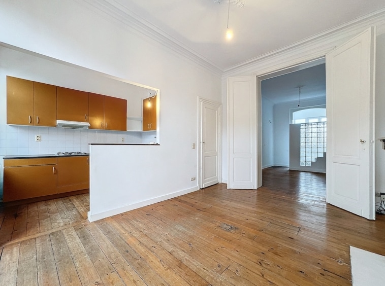 Salon et cuisine ouverts, lumineux avec grandes fenêtres, murs blancs, sols en bois, ambiance moderne et chaleureuse.