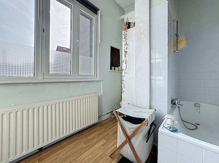 Salle de bain lumineuse avec baignoire blanche, fenêtres, plancher en bois et rangement pratique.
