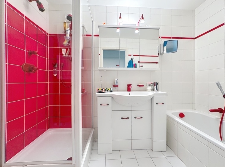 Salle de bain contemporaine avec accents rouges, douche en verre, baignoire blanche et éclairage chaleureux.