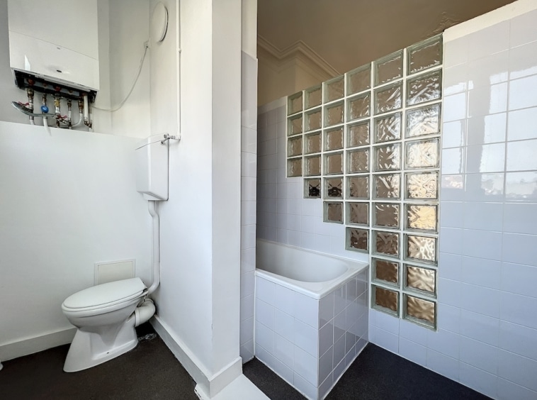 Salle de bain moderne compacte avec douche en blocs de verre, baignoire blanche, murs lumineux, sol gris foncé.