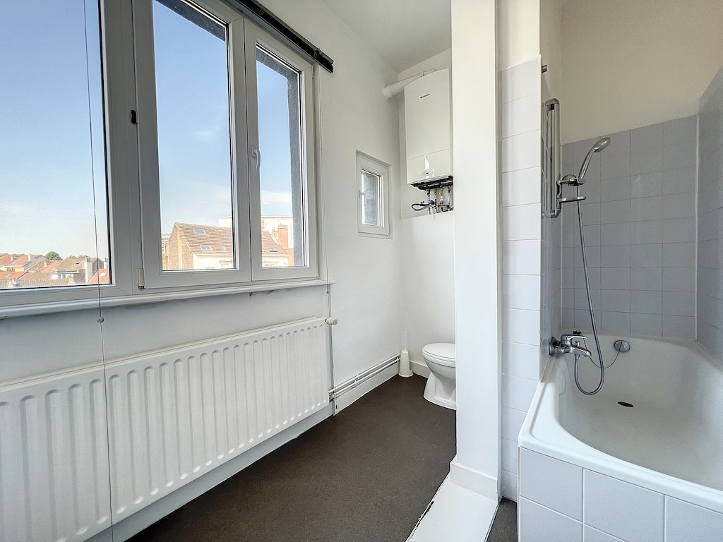 Salle de bain moderne lumineuse avec baignoire, douche, WC, fenêtre, radiateur et vue extérieure.