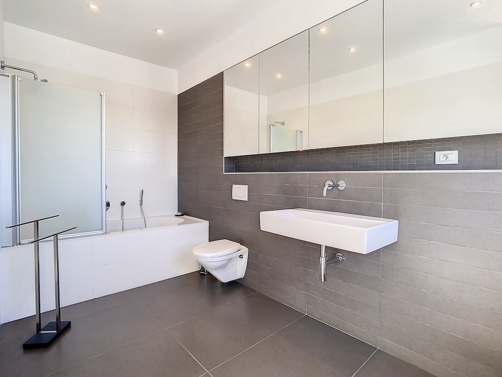 Salle de bain moderne, épurée, lumineuse avec douche en verre, baignoire, lavabo mural et miroir.