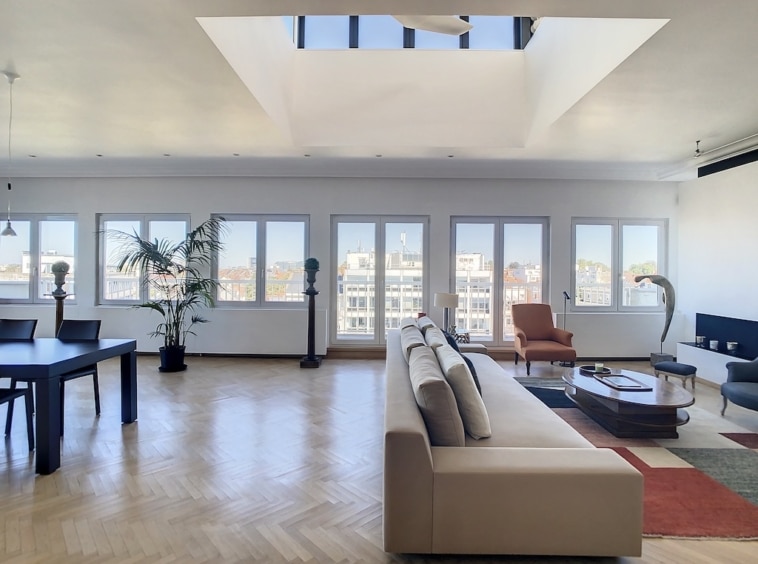 Salon moderne lumineux avec grandes fenêtres, vue urbaine, mobilier élégant et touches végétales.