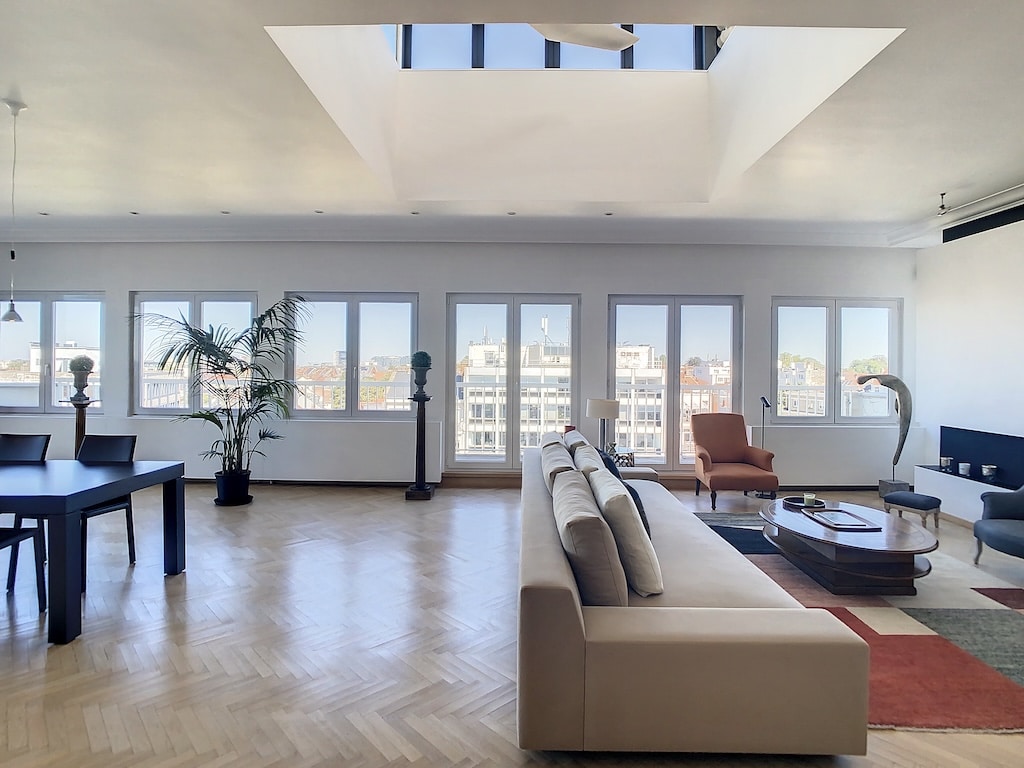 Salon moderne lumineux avec grandes fenêtres, vue urbaine, mobilier élégant et touches végétales.