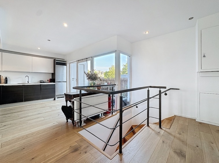 Salon contemporain lumineux avec cuisine moderne, escalier en métal noir, vue sur jardin verdoyant.