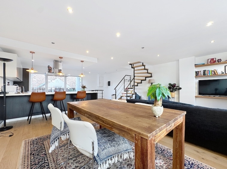Salon contemporain lumineux avec cuisine ouverte, mobilier blanc et bois, grand canapé noir, escalier moderne.
