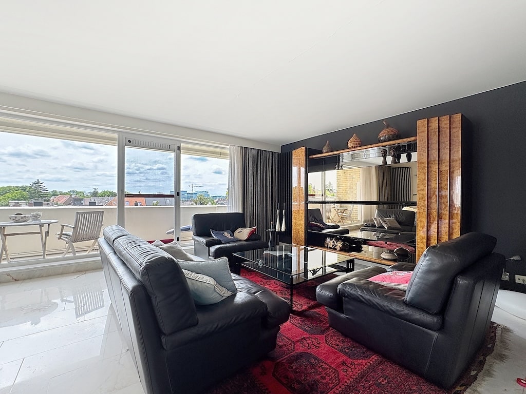 Salon lumineux moderne avec fenêtres, mobilier élégant en cuir noir, tapis rouge, et meuble en bois.