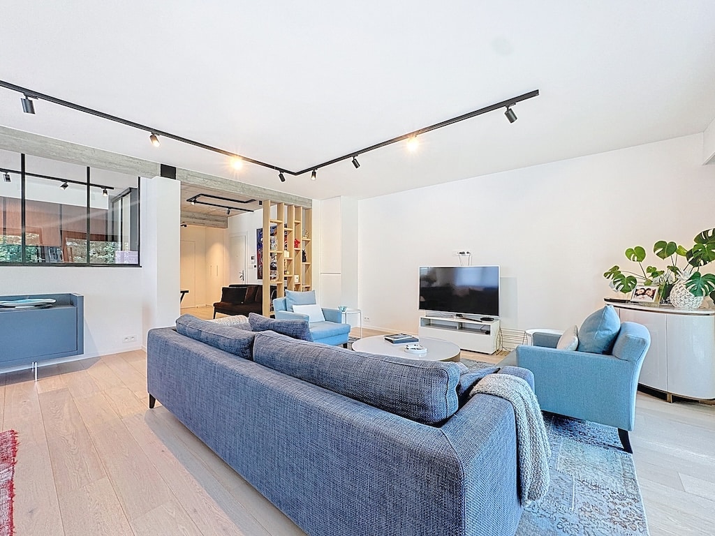 Salon moderne lumineux avec canapé bleu, fauteuils, mobilier épuré, grande fenêtre, vue cuisine ouverte.