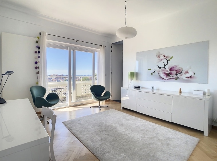Salon lumineux moderne avec mobilier épuré, vue extérieure, chaises teal, œuvre florale, éclairage élégant.