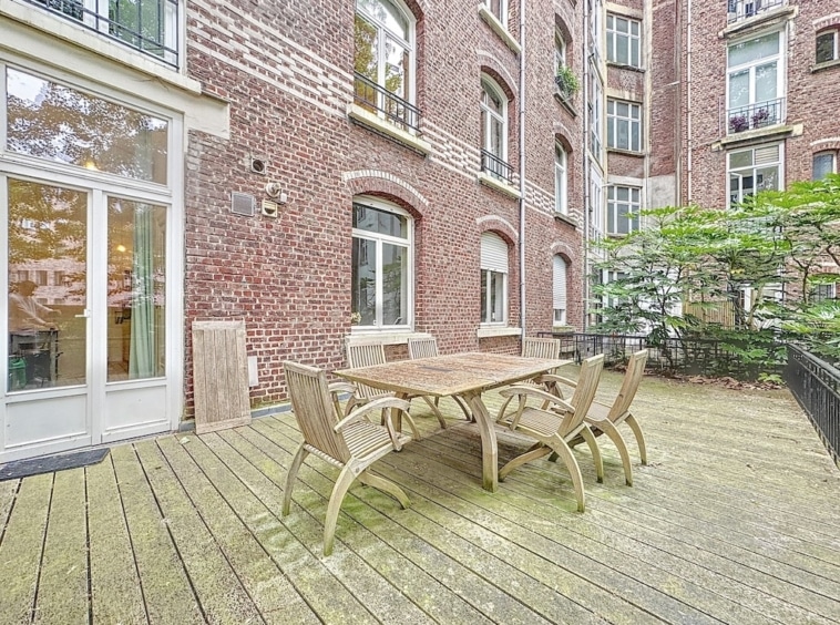 Terrasse en bois rustique avec mobilier élégant, façade en briques anciennes et ambiance chaleureuse.