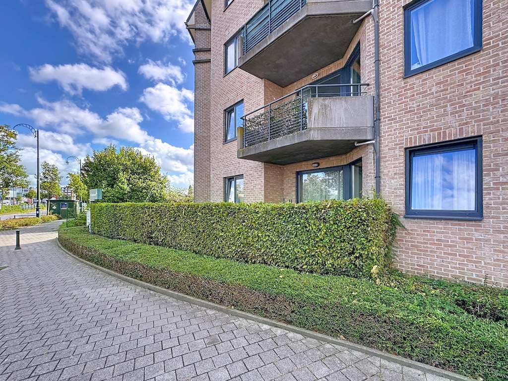 Immeuble moderne en briques avec balcons en verre, jardin urbain, chemin pavé et ciel bleu.
