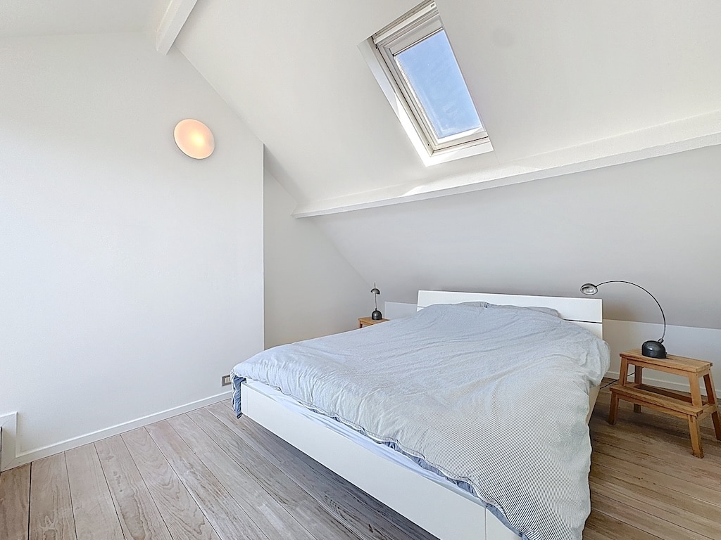 Chambre attic lumineuse minimaliste avec lit moderne, plancher en bois clair et lumière naturelle.