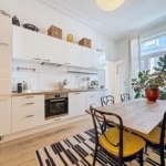 Cuisine moderne lumineuse avec mobilier blanc, table en bois et touches de couleur chaleureuses.